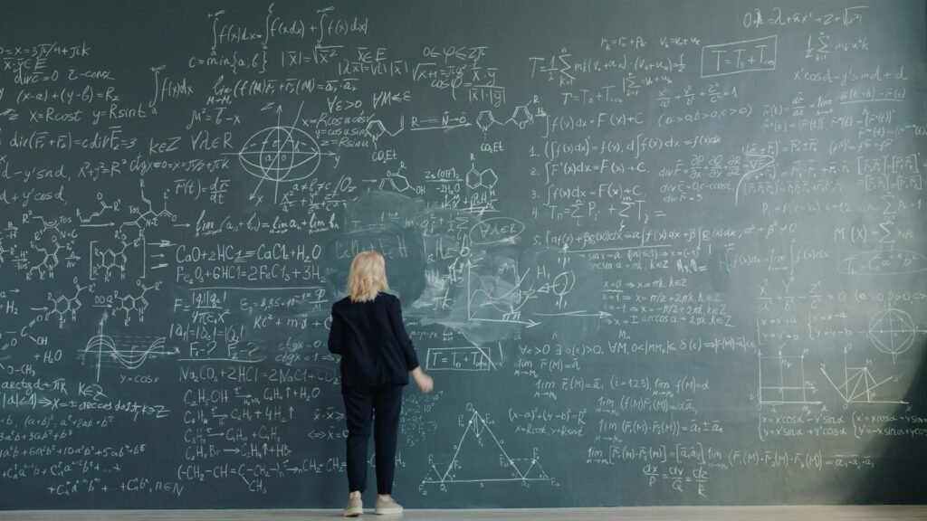 A teacher facing a wide chalkboard covered in mathematics and science working