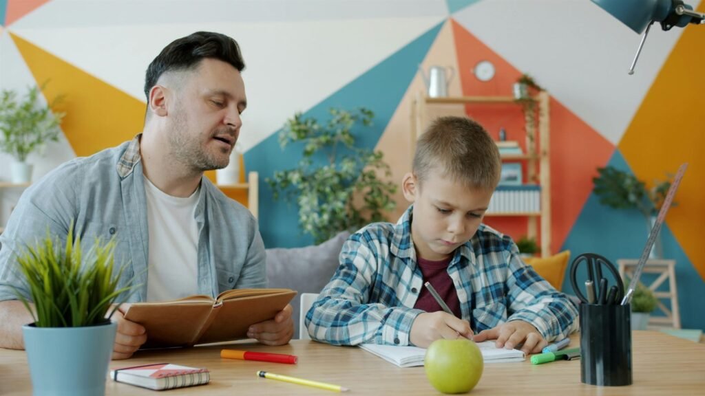 A private tutor sitting beside a young boy as he writes in his exercise book at home